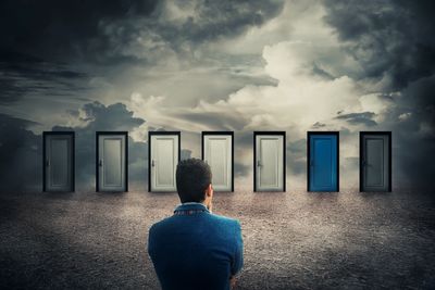 Man in blue jacket looking at seven doors with clouds behind
