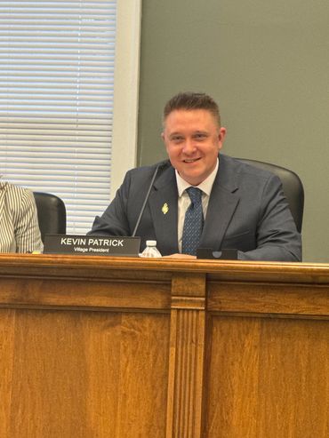 Kevin Patrick, Village President, seated at a council meeting desk.