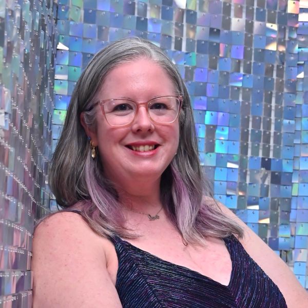 Smiling woman with glasses and purple streaks in her hair against a shiny, mosaic background.