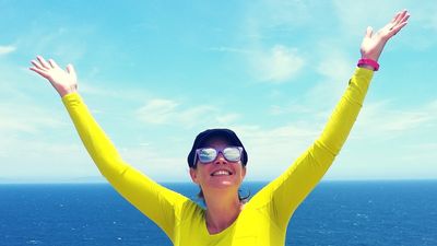 Shows a woman in front of the ocean holding her arms in a victorious gesture