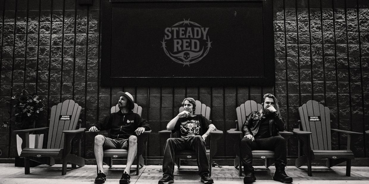 Three men sitting in chairs under a 'Steady Red' sign, black and white photo.