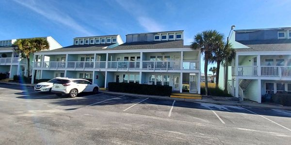 Two-story beachside apartments with parked cars and palm trees under a clear blue sky.