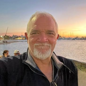 Smiling man taking a selfie by the water at sunset with boats in the background.