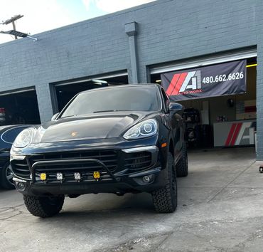Black Porsche SUV with off-road lights parked outside an auto repair shop.