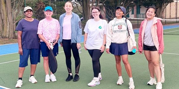 Six women standing and smiling on a tennis court, dressed in sporty casual wear.