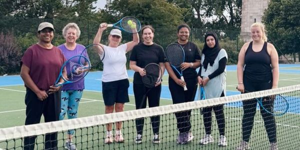 Seven women posing with tennis rackets on a court, smiling and ready to play.