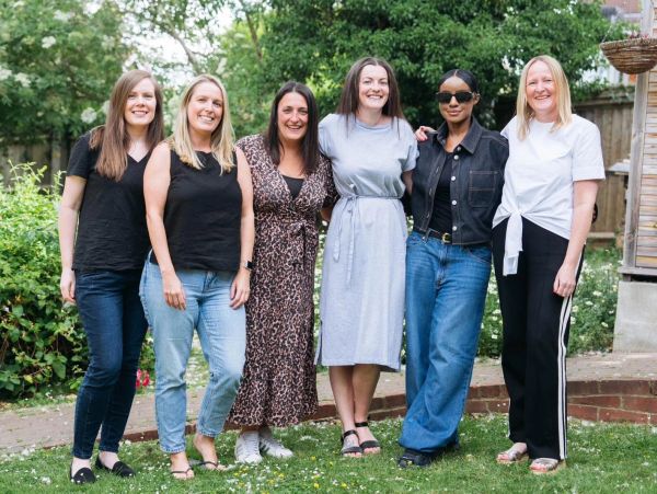 Six women standing together outdoors, smiling and posing for a group photo.