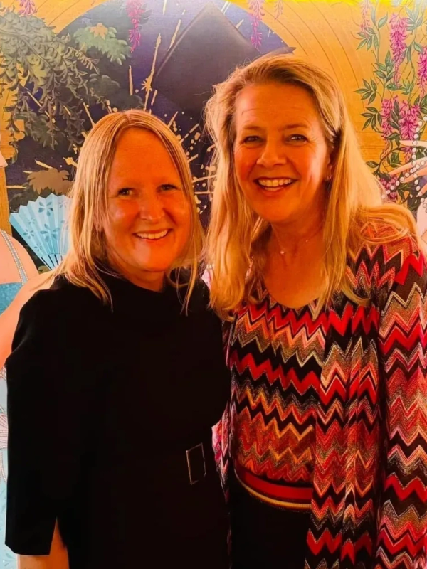 Two smiling women pose in front of a colorful, artistic mural.