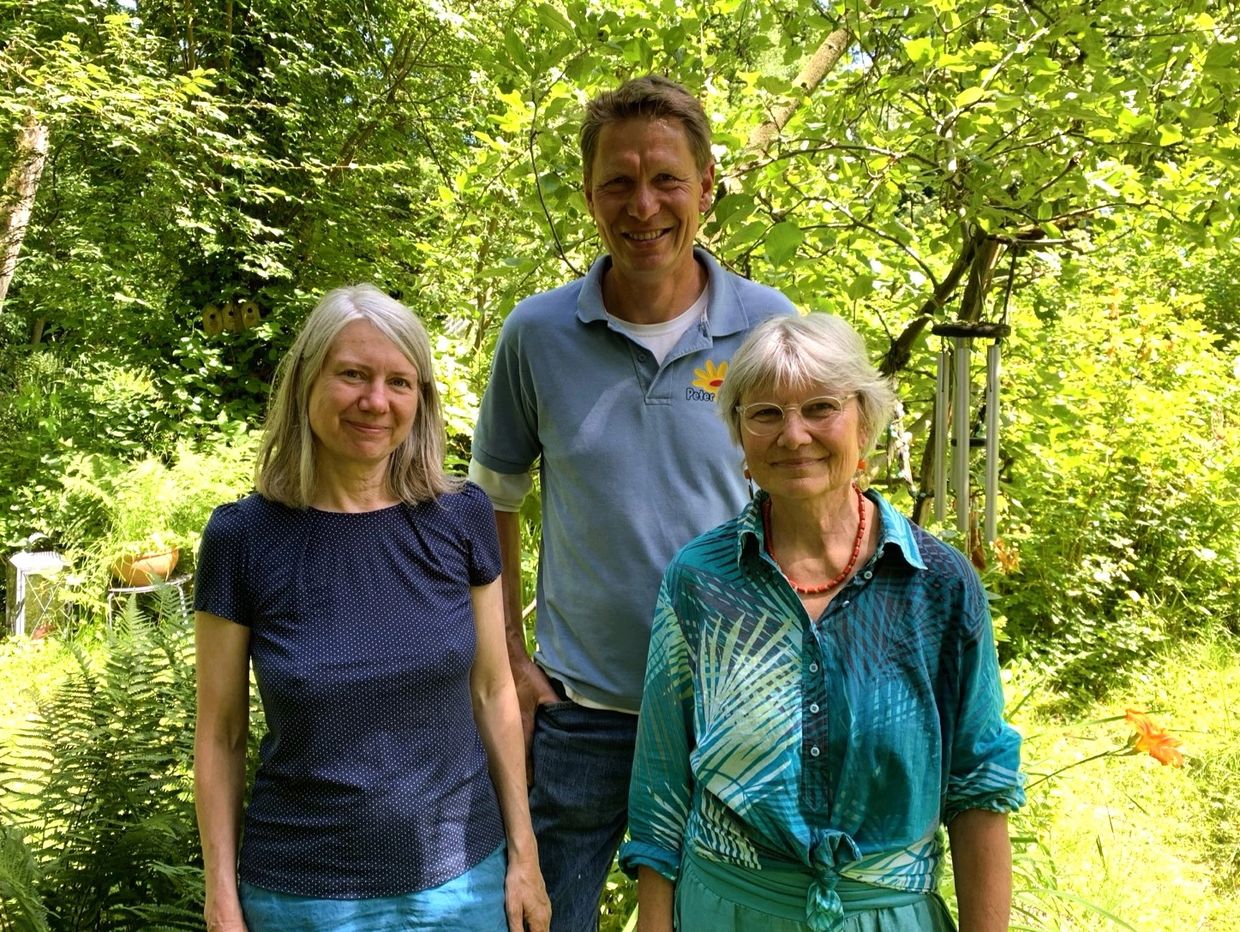 Drei lächelnde Menschen im Garten vor grüner Natur.