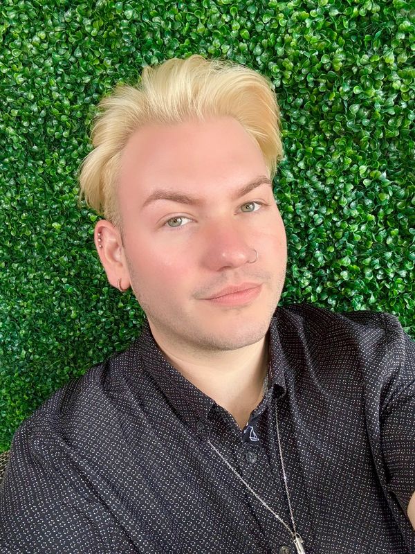 Person with blonde hair and earrings posing against a green leafy background.