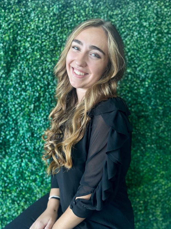 Young woman with long curly hair smiling against a green leafy background.