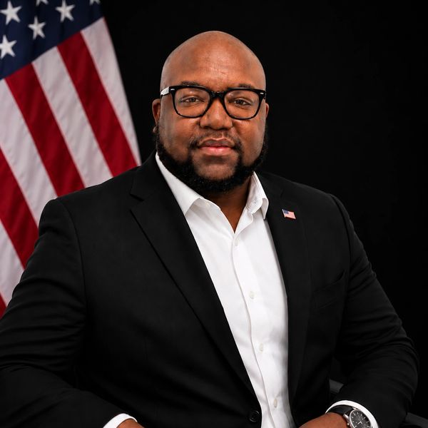 Man in a suit with an American flag pin and flag backdrop.