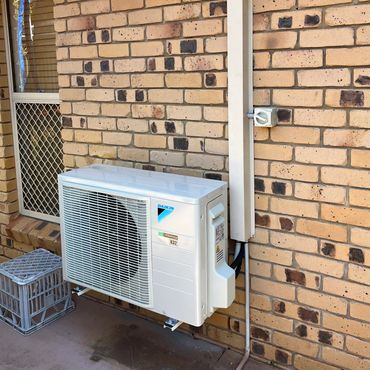 Outdoor air conditioning unit mounted on a brick wall near a window and a plastic crate.