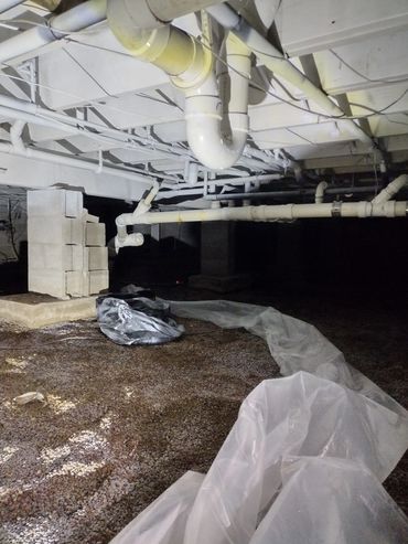 Dimly lit crawl space with pipes and plastic sheeting on gravel floor.