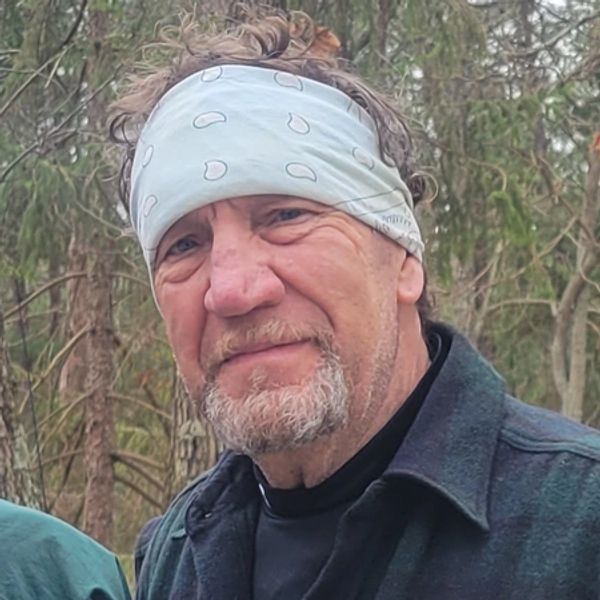 Man with a white bandana and beard outdoors among trees.