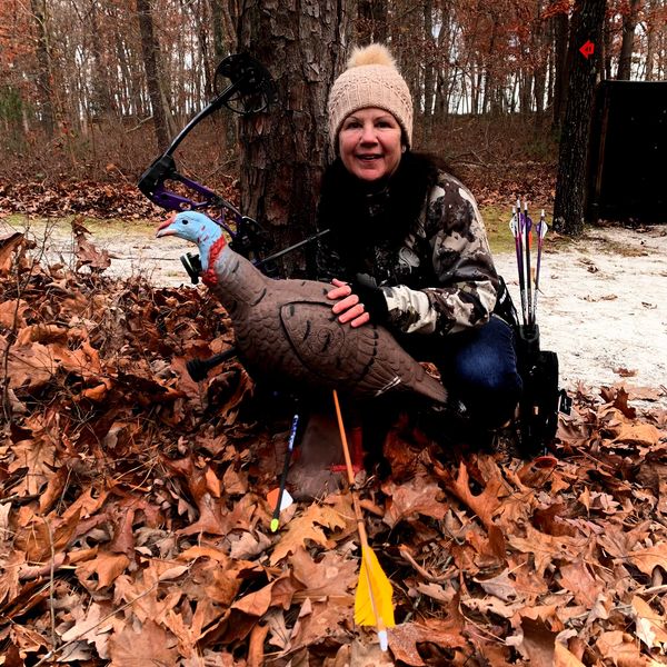 Woman dressed for hunting poses with a bow and a turkey target in fallen leaves.