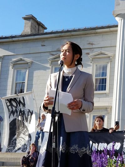 Rev. Joan Javier-Duval delivers speech outside statehouse wearing her collar and light gray blazer.