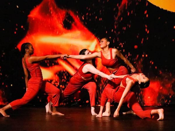 Four contemporary dancers in burnt orange costumes tangled up and holding hands onstage