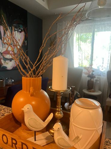 Decorative orange vase with branches, white birds, and a candle holder on a wooden tray.