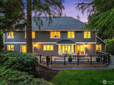 A cozy two-story house with warm lights and a spacious deck surrounded by lush greenery at dusk.