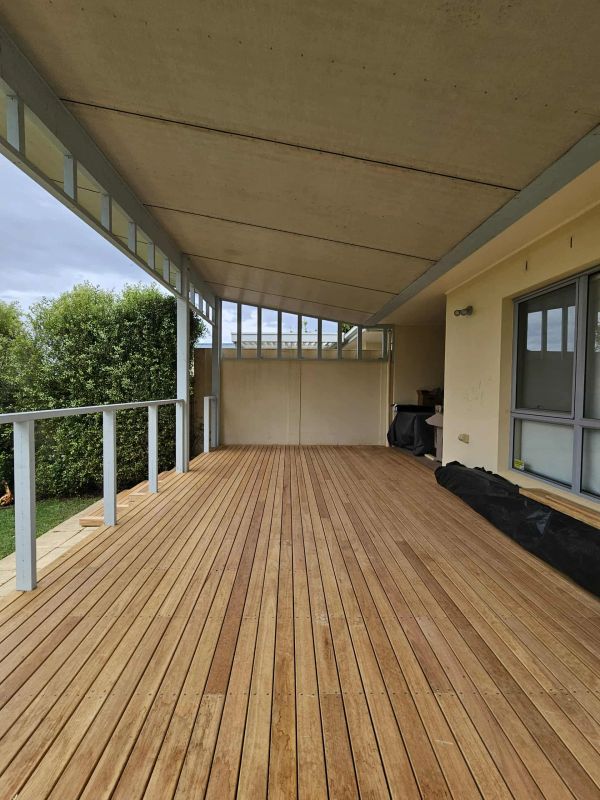 Empty covered wooden deck with railing and green bushes.