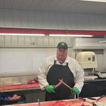 Man in black apron and green gloves holding knives in a butcher shop.