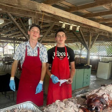 Two young men in red aprons preparing meat at an outdoor kitchen.