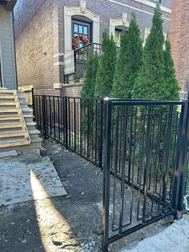 Newly installed black metal fence with green shrubs and a brick building.