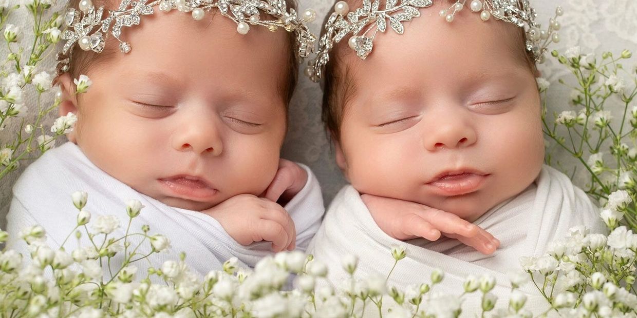 Sleeping newborn twins adorned with delicate pearl and crystal headbands among white flowers.