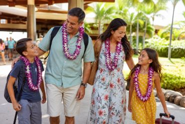 Family of four wearing fresh orchid leis and walking with luggage after arriving in Hawaii