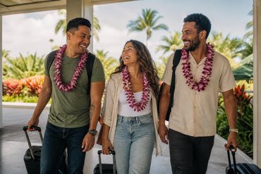 Three friends wearing fresh orchid leis and carrying luggage after arriving in Hawaii