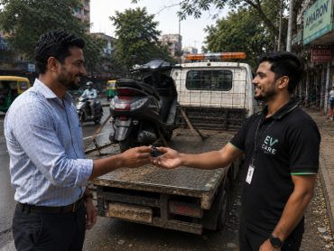 Two men exchanging scooter keys beside a tow truck on a city street.