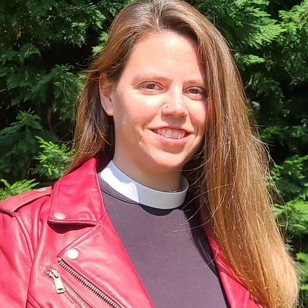 Woman with long hair wearing a red jacket and clerical collar, smiling outdoors.