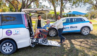 Caregivers assisting a man in a wheelchair into a vehicle.