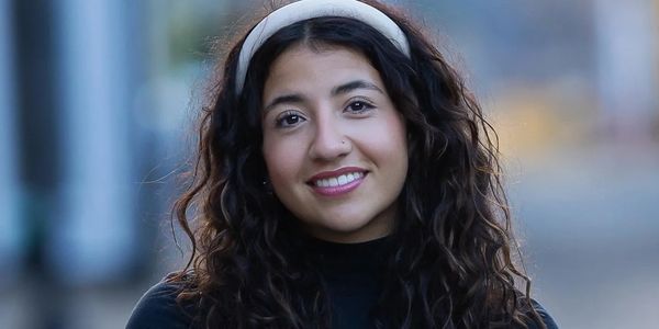 Young woman with curly hair and headband smiling while sitting on a chair outdoors.
