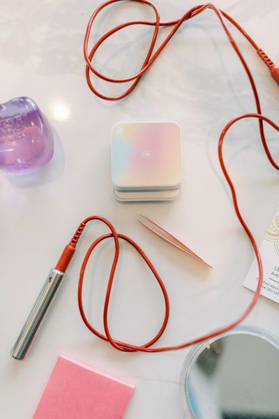 Beauty tools including tweezers, a small mirror, and a device with red cords on a table.