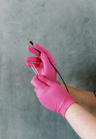Hands in pink gloves holding an electrode and tweezers against a gray background.