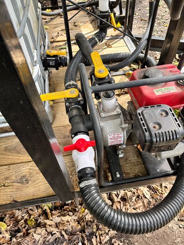 A gas-powered water pump with hoses and valves on a wooden platform outdoors.