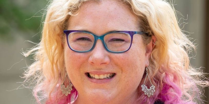 Smiling woman with blonde and pink hair wearing glasses and a black polka dot top.