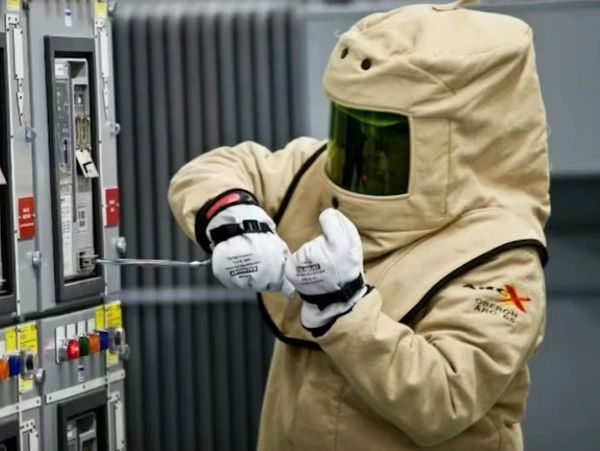 A technician in a full-body arc flash suit uses a tool to work on an electrical panel.
