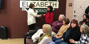 Two women shake hands in front of a group in a community room.