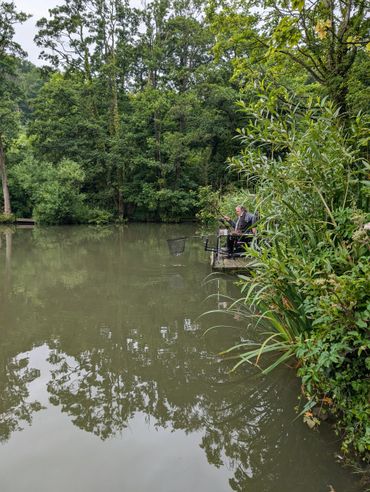 Man fishing quietly on a wooden platform by a serene, tree-lined lake.