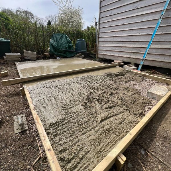 Fresh concrete slab being poured and leveled next to a wooden shed.