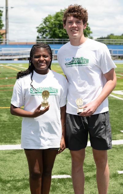 Two teens holding trophies on a sports field wearing matching charity event shirts.