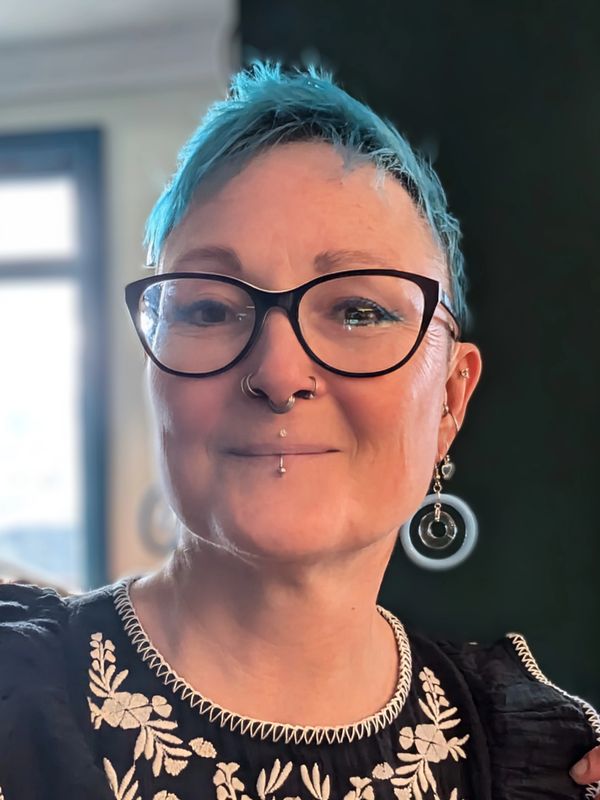 Smiling person with blue hair, glasses, and piercings.