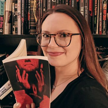 Woman with glasses holding a book in front of a bookshelf.