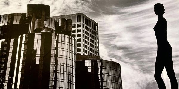 Silhouette of a statue against modern buildings and a cloudy sky.