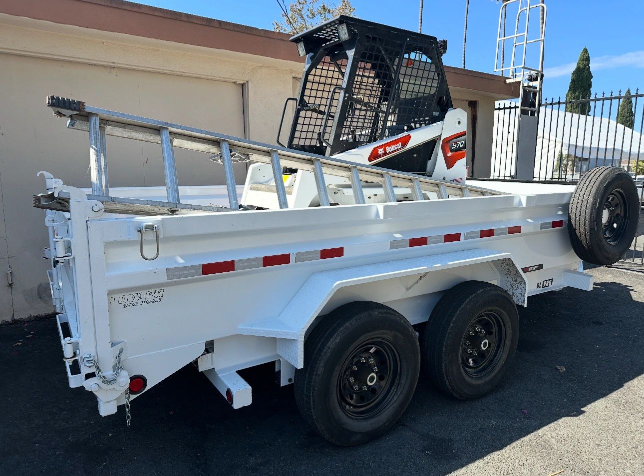 White utility trailer with ladder and Bobcat skid steer inside, parked outdoors.