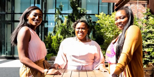 Three women smiling around a wooden table outdoors in sunlight.