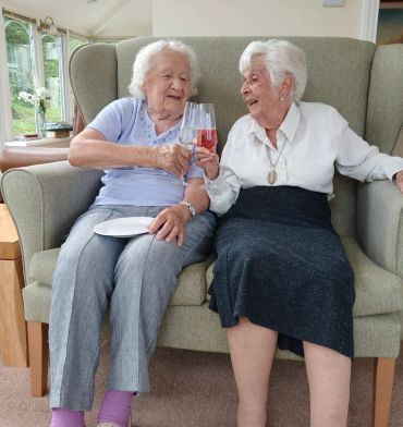 Two elderly women cheerfully toast while sitting on a couch.